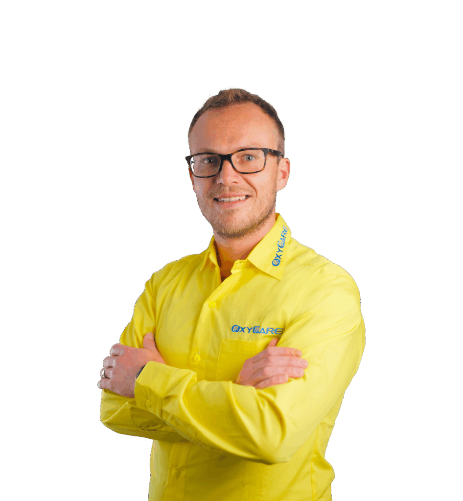 Foto von Pierre Lange, Geschäftsführer von OxyCare. Er steht mit vor der Brust verschränkten Armen da, trägt ein gelbes Hemd mit dem OxyCare Logo und schaut seriös in die Kamera.