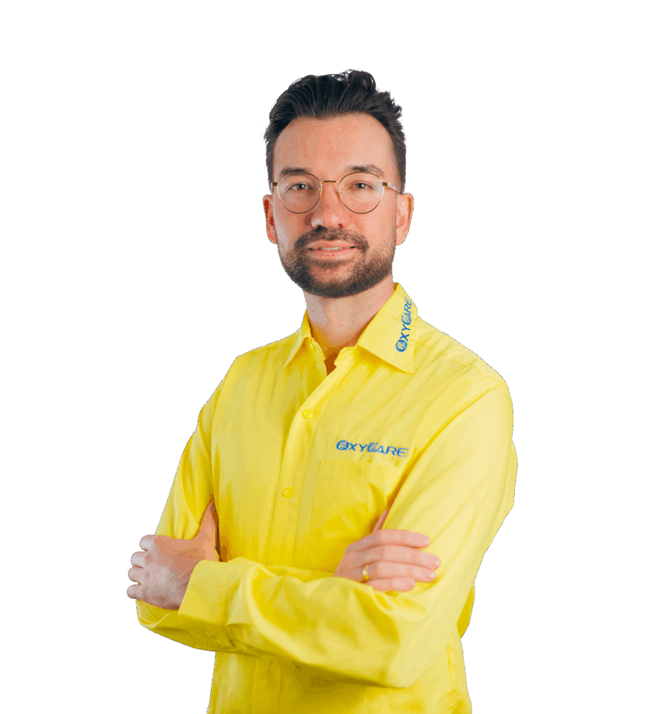 Foto von René Drescher, Geschäftsführer von OxyCare. Er steht mit vor der Brust verschränkten Armen da, trägt ein gelbes Hemd mit dem OxyCare Logo und schaut seriös in die Kamera.